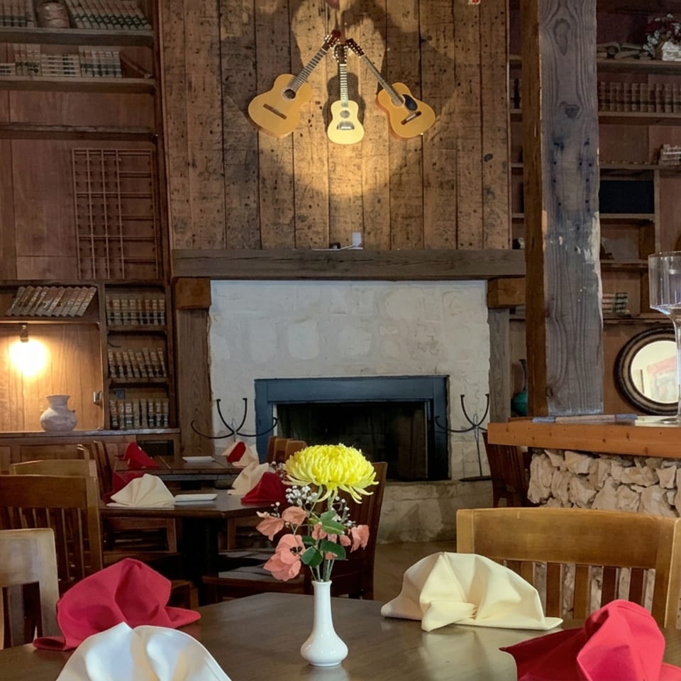 Rustic restaurant dining room with wooden tables, colorful napkins, floral centerpiece, stone fireplace, guitars, and wine glass.
