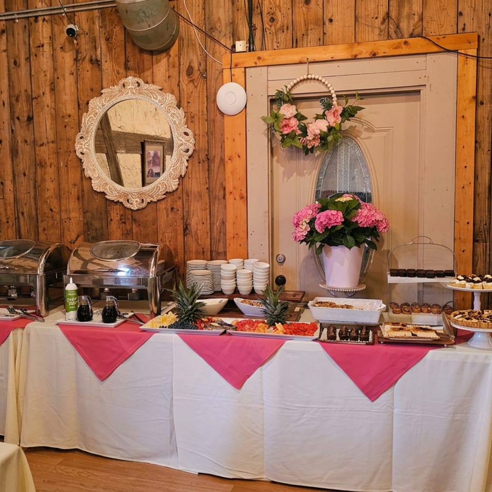 Buffet table with chafing dishes, desserts, fruit, floral decorations, and pink-and-white table linens in a rustic wood-paneled room.