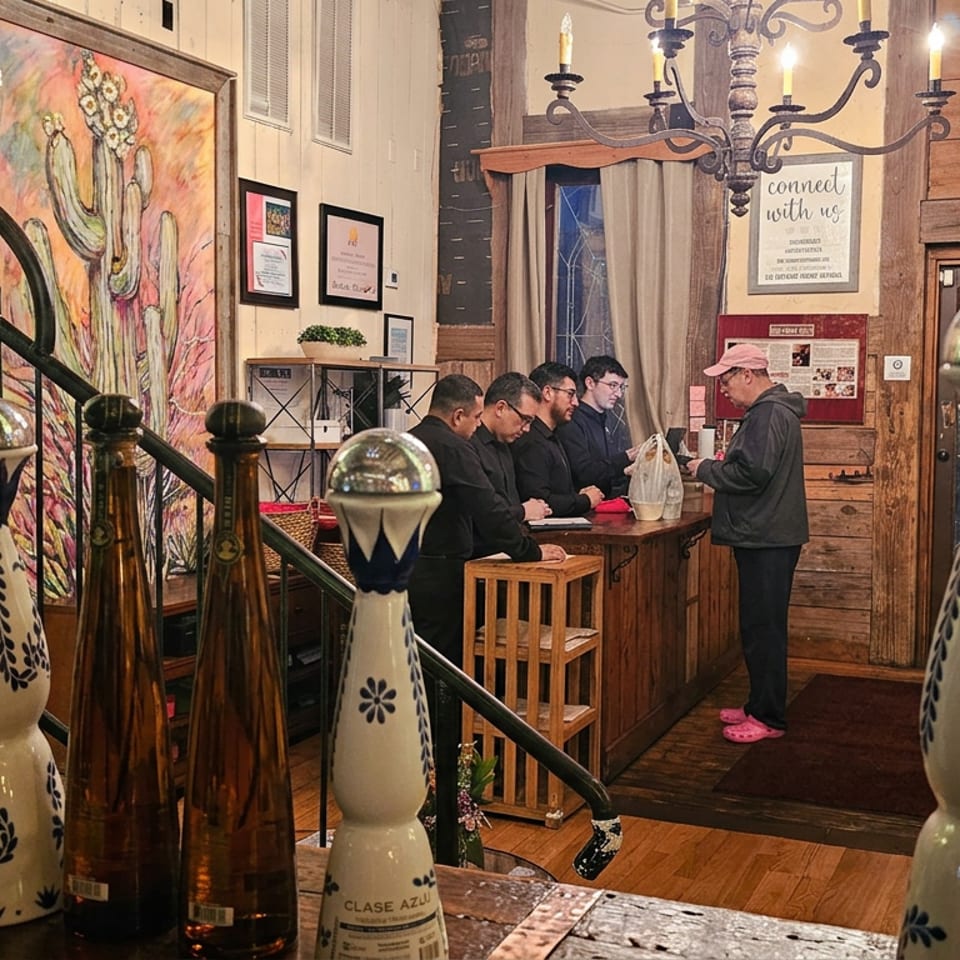 Rustic restaurant interior with hanging glasses, tequila bottles, chandelier, artwork, and guests gathered at the host counter.