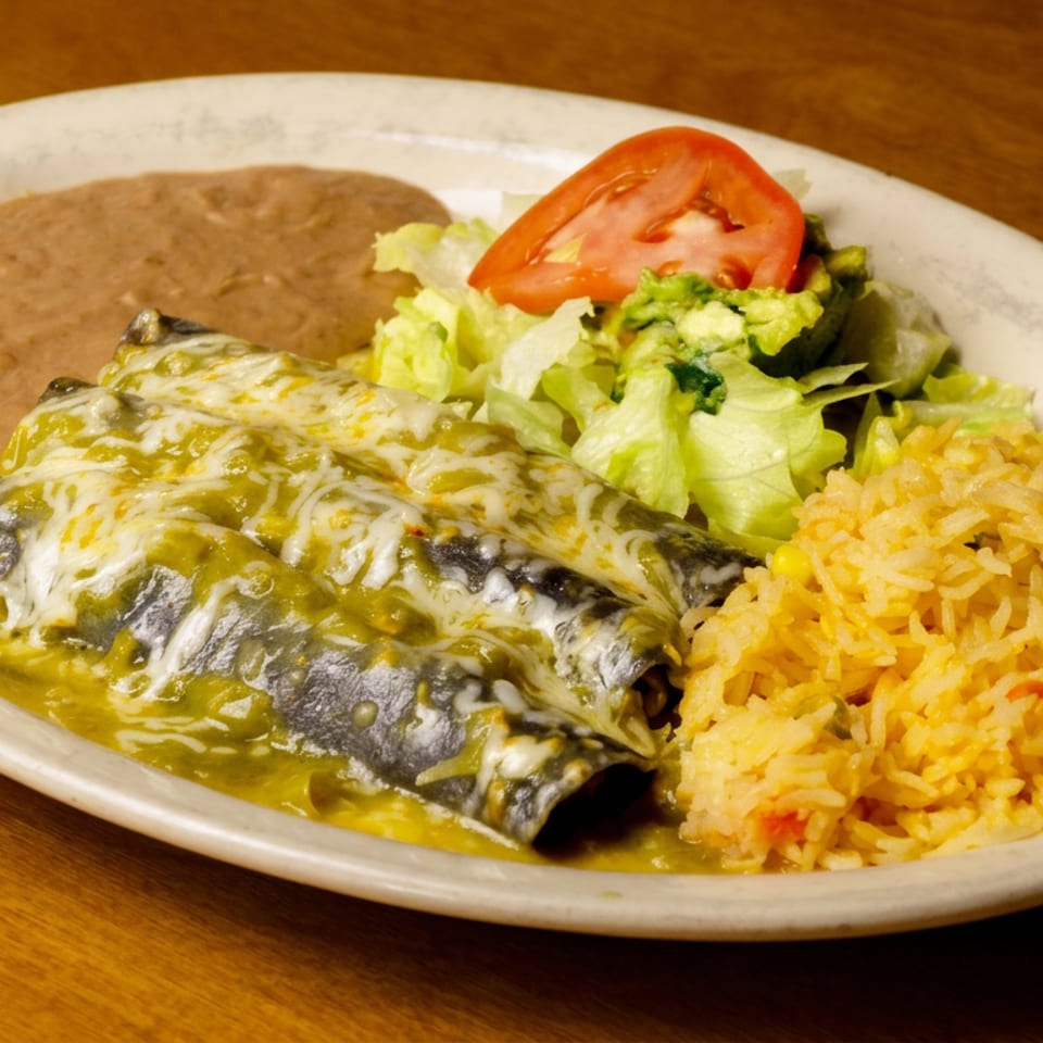 Mexican entrée with cheese-covered green enchiladas, refried beans, rice, lettuce salad, guacamole, and tomato on a white plate.