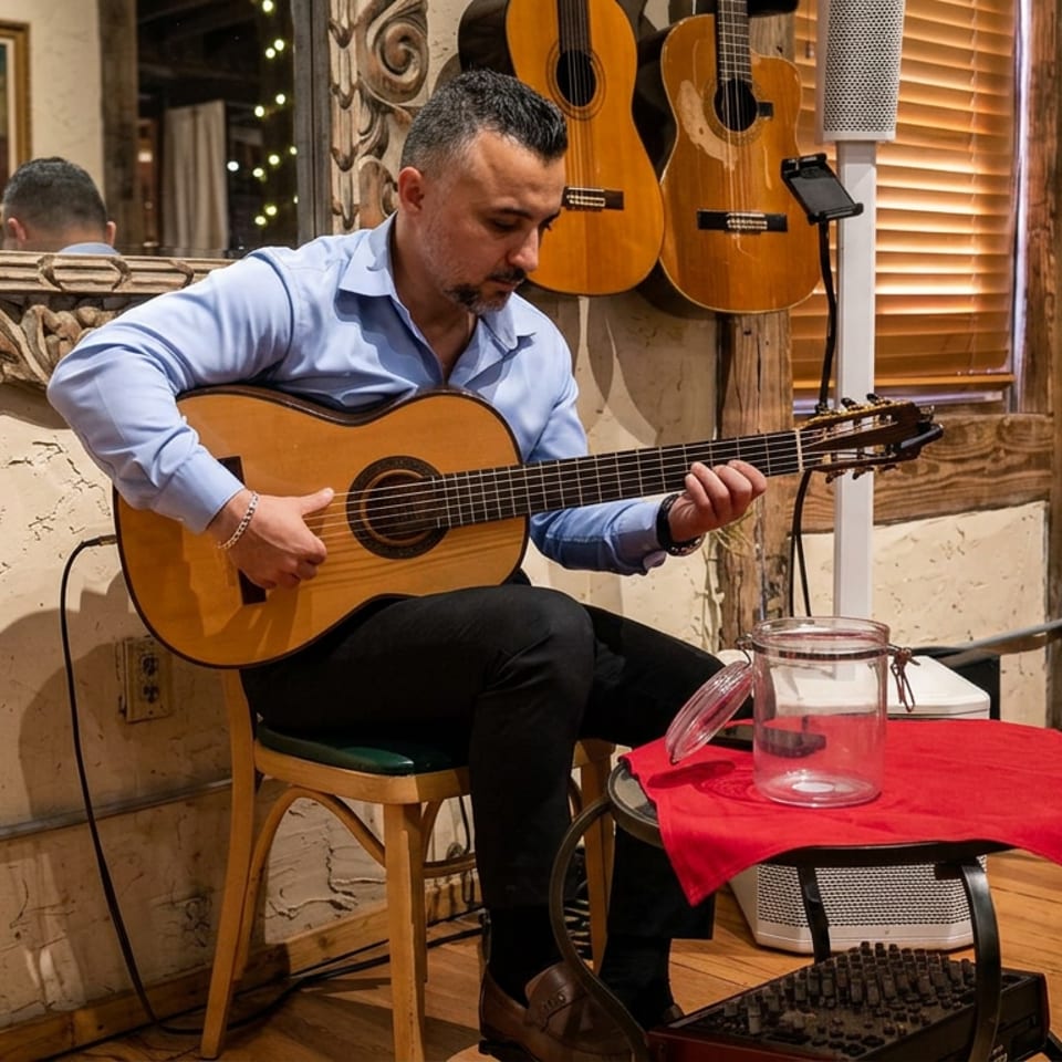 Musician plays acoustic guitar in a warmly decorated restaurant, with hanging guitars, tables, and a seated diner nearby.