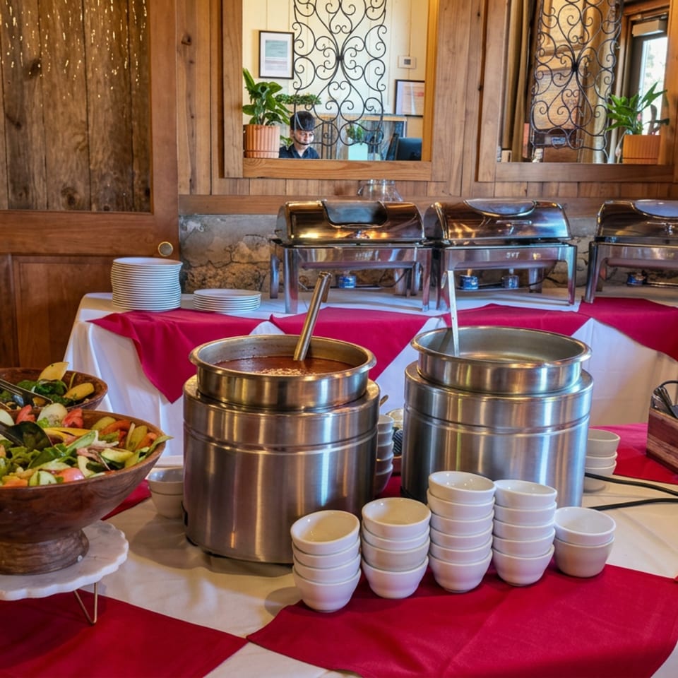 Buffet station in a rustic restaurant featuring salad bowls, soup kettles, chafing dishes, stacked plates, and serving utensils.