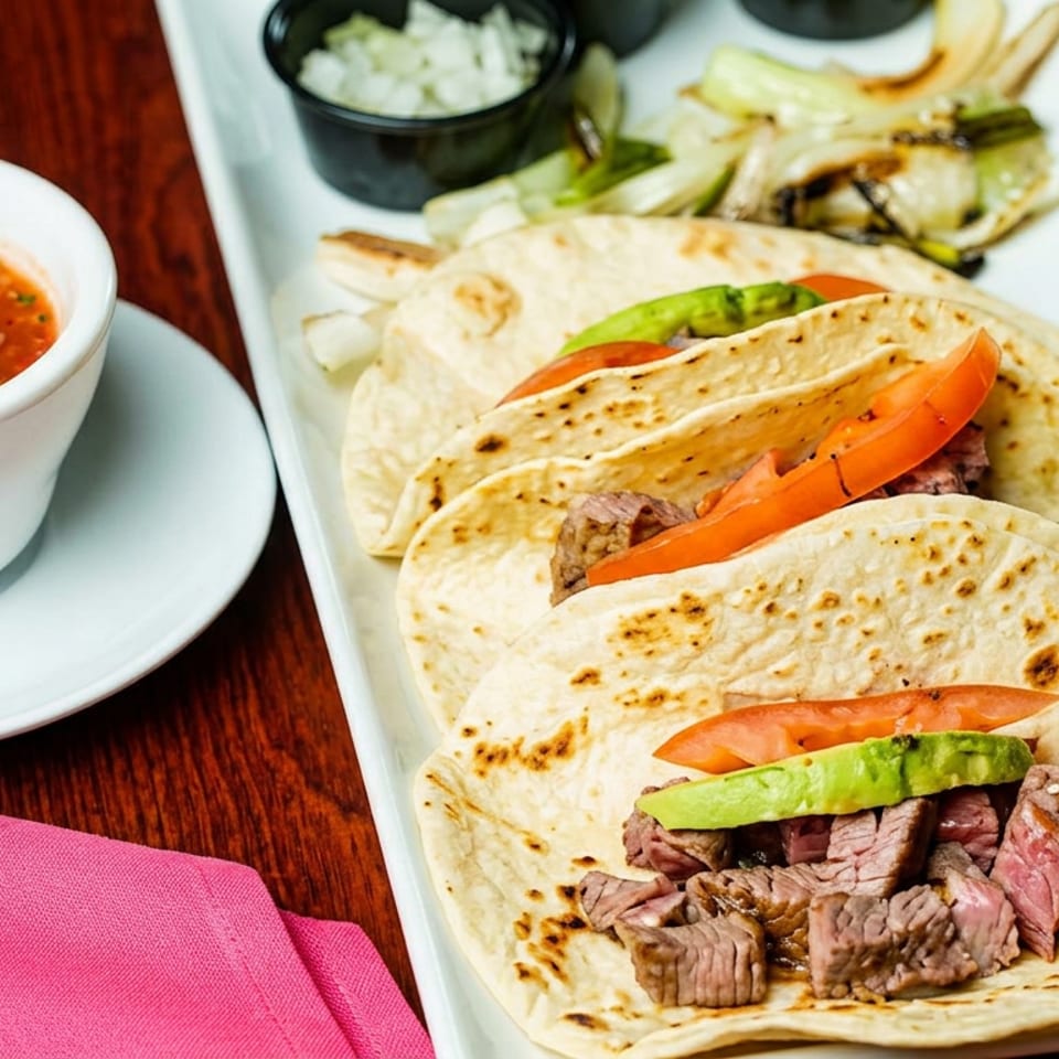 Three grilled steak tacos with avocado and tomatoes served alongside salsa, rice, and fries on a white platter.