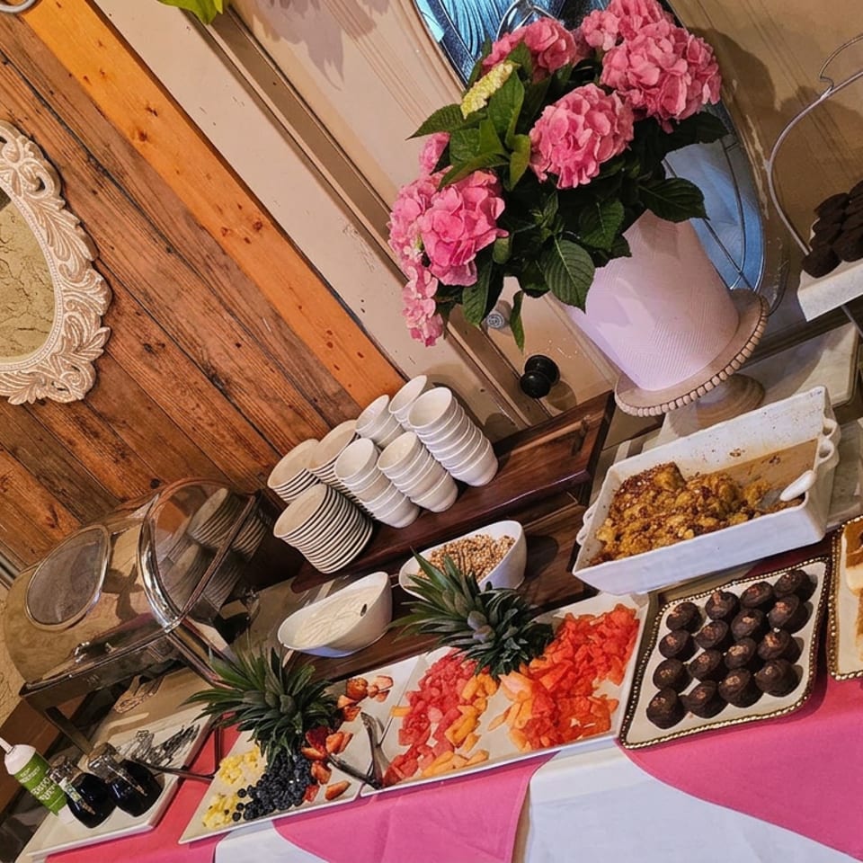 Dessert buffet displays pastries, chocolate treats, waffles, fresh fruit, and pink flowers in a rustic restaurant setting.