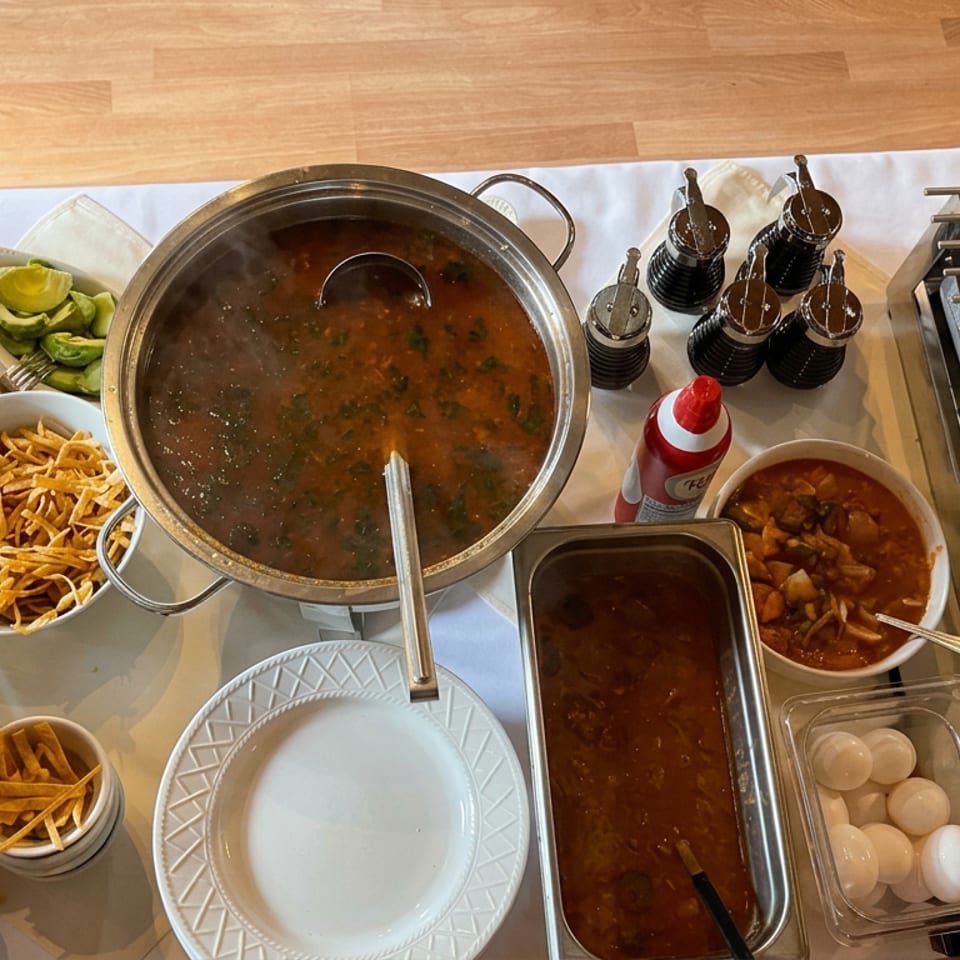 Overhead view of a restaurant buffet with simmering soup, tortilla strips, avocados, sauces, eggs, and cooking equipment.