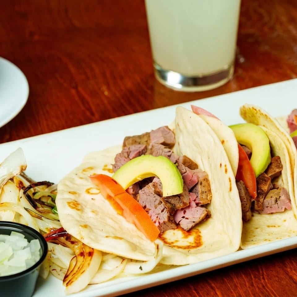 Platter of soft steak tacos topped with avocado and tomato, served with grilled onions, sauces, salsa, and a beverage.