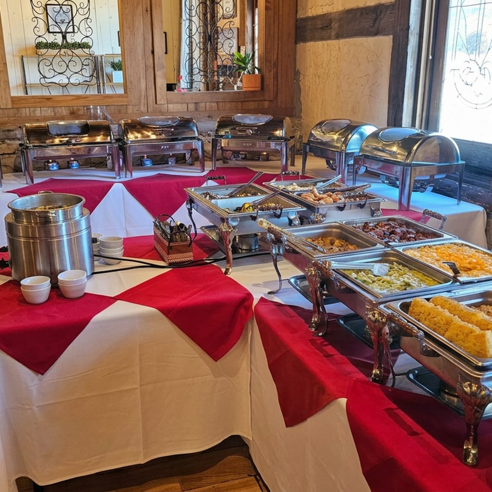 Buffet spread with chafing dishes, assorted hot foods, red-and-white tablecloths, and beverage service in a rustic restaurant dining room.